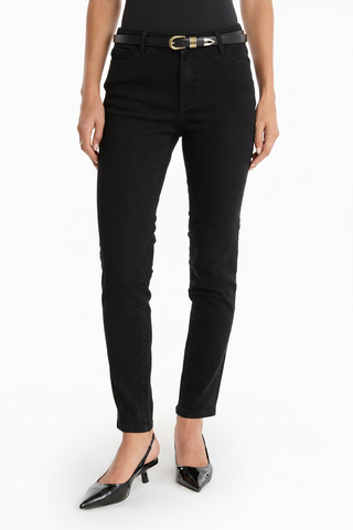A person wearing the Layla Skinny Jean - Black, a black belt with a gold buckle, a black top, and black pointed-toe heeled shoes stands against a white background.