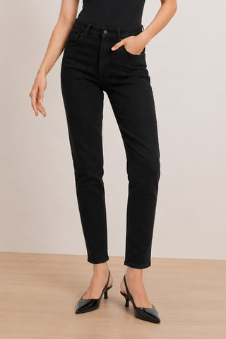 Wearing the Layla Skinny Jean in Black, which features a flattering fit and comfortable stretch denim, a person stands on a wooden floor with one hand in their pocket and the other arm relaxed by their side. They complete the look with a black top and black heeled shoes, making this outfit both stylish and versatile for any occasion.