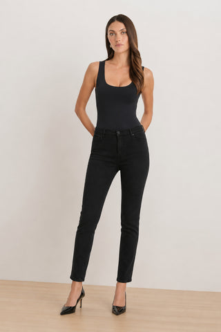 A woman wearing a fitted black tank top, the Layla Skinny Jean - Black with a flattering fit, and black pointed-toe heels stands on a light wood floor against a plain off-white background, with her hands behind her back.