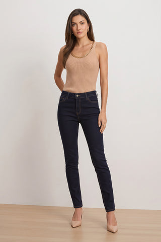 A woman stands on a wooden floor wearing a beige sleeveless top, the Layla Skinny Jean in Indigo, and beige heels. She faces forward with her hands behind her back against a plain light background.