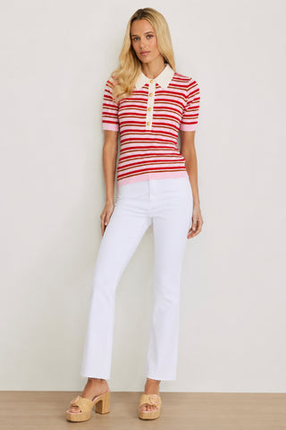 A woman with long blonde hair poses casually against a light background, wearing the Leya Polo Shirt - Summer Multi in red and white stripes, styled with crisp white pants and tan platform shoes.