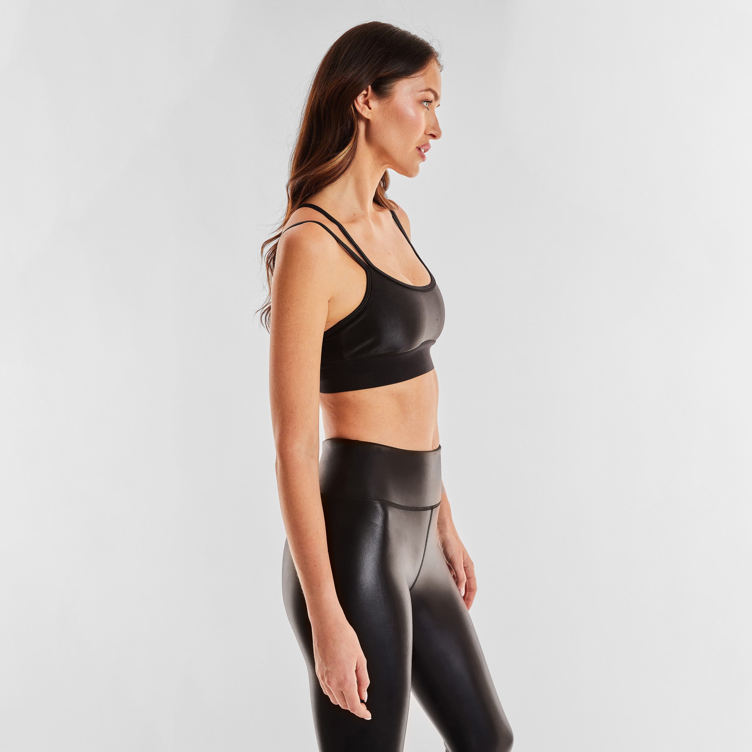 Side view of woman wearing black gloss liquid sports bra featuring a scoop neckline and an alluring strappy back with a supportive elastic band