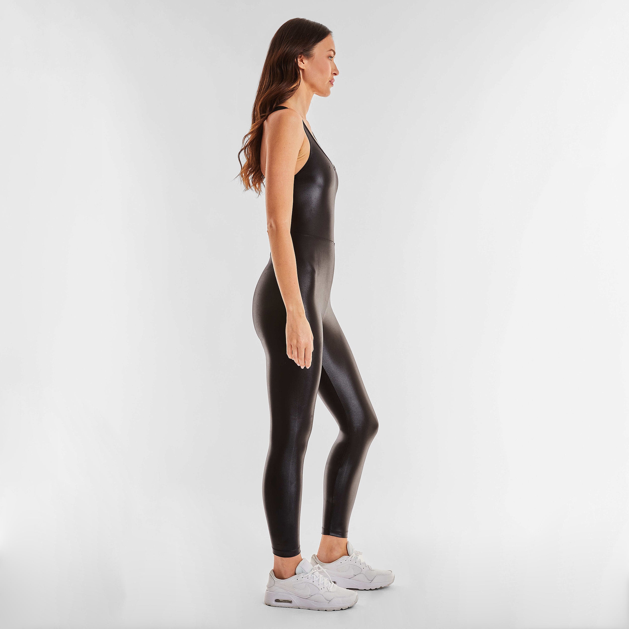 Side view of woman wearing black jumpsuit designed with a figure-flattering waistline and strappy back. Designed with our iconic Liquid shine fabric.