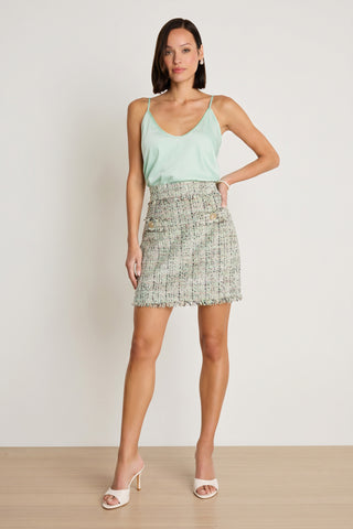 A woman poses against a plain background wearing a light green sleeveless top, white open-toe heels, and the Lisette Tweed Skirt - Teal Multi, a high-waisted luxury mini skirt with a frayed hem. She stands with one hand on her hip, facing the camera.