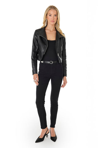 A woman with long blonde hair, minimal jewelry, and a confident expression poses against a white background wearing the Katalina Moto Jacket - Black with an asymmetrical silhouette, black top, skinny jeans, and pointed-toe heels.