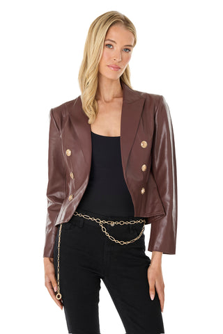 A woman with long blonde hair wears the Gabrielle Leather Double Breasted Blazer in Espresso, featuring gold buttons, over a black top and pants with a gold chain belt. She stands against a white background.