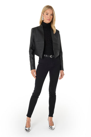 A woman stands confidently against a white background wearing the Yasmin Leather Jacket - Black, cropped with an asymmetrical hem, paired with a black turtleneck, skinny jeans, silver heels, black belt, and statement earrings.