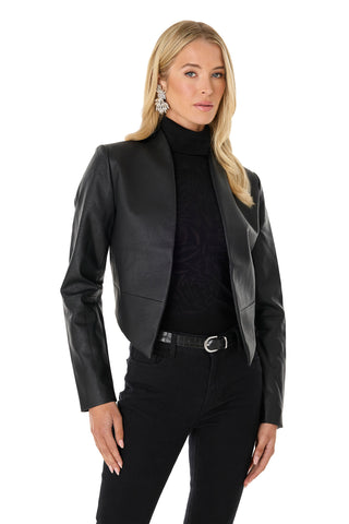 A woman with long blonde hair wears the Yasmin Leather Jacket - Black, featuring an asymmetrical hem, over a black turtleneck and pants. She faces forward and accessorizes with large silver earrings.