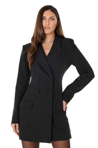 A woman with long brown hair wears the Kira Blazer Dress - Black, featuring power shoulders and flap pockets. She stands facing forward with one hand on her hip against a plain white background.