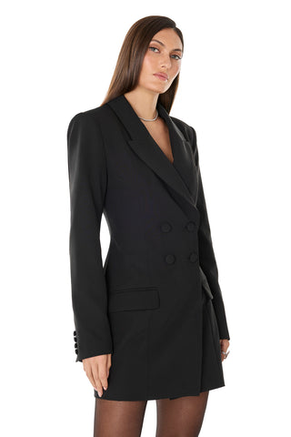 A woman with straight brown hair models the Kira Blazer Dress - Black, a suiting-style dress with power shoulders, long sleeves, black buttons, and front pockets, standing against a plain white background.