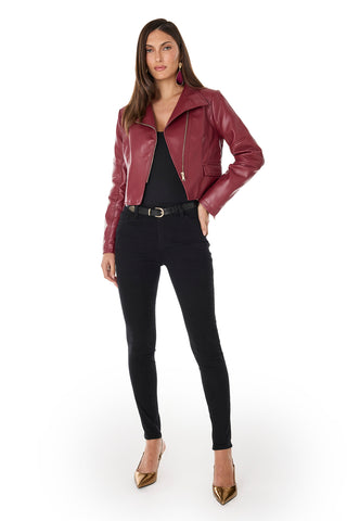 A woman faces forward in a chic Katalina Moto Jacket - Merlot over a black top and skinny jeans, with gold pointed-toe heels. Her straight brown hair and one hand in her pocket accentuate the jacket’s asymmetrical silhouette on a white background.
