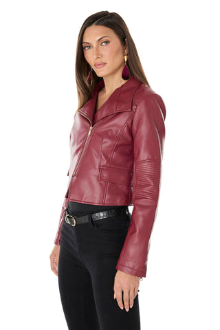 A woman with long brown hair wears the Katalina Moto Jacket - Merlot, styled with black pants and red earrings. She stands angled to the camera against a white background, her expression neutral.