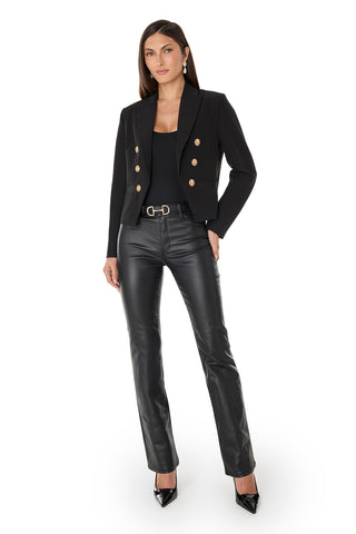 Woman with long, straight hair and earrings stands against a white background wearing the Gabrielle Double Breasted Blazer - Black with gold buttons, a black top, black leather pants, and black pointed-toe heels.