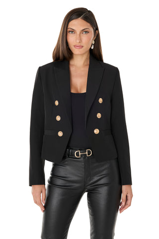 A woman with straight brown hair and an earring stands facing forward against a white background, wearing the Gabrielle Double Breasted Blazer - Black with gold buttons, a black top, and black leather pants.