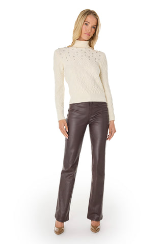 A woman stands against a white background wearing the Cara Crystal Sweater - Pearl, featuring cable-knit details, paired with dark brown leather pants and beige heels—an effortless winter layering choice.