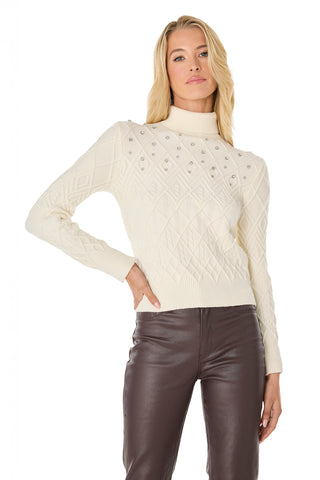 A woman wears the Cara Crystal Sweater - Pearl, featuring a diamond pattern and high collar, ideal for winter layering. She pairs it with high-waisted dark brown leather pants and poses confidently with one hand on her hip.