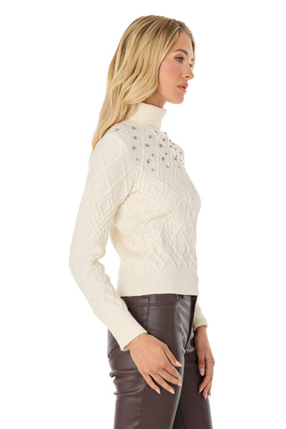 A woman with long blonde hair models the Cara Crystal Sweater - Pearl, a white cable-knit turtleneck with chest button details, paired with dark brown leather pants and standing in profile against a white background.