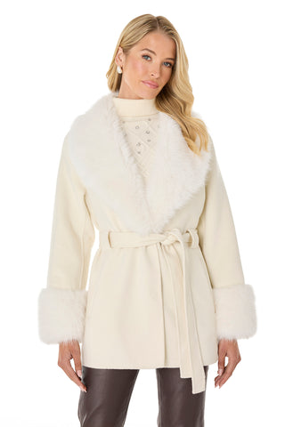 A woman wears the Isabel Fur Trim Coat in white—featuring a belted design and faux fur at the collar and cuffs—paired with a cream turtleneck sweater and dark pants, standing against a plain white background.
