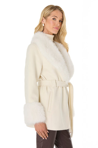 A woman with long blonde hair poses against a white background wearing the Isabel Fur Trim Coat in white, featuring a timeless silhouette and belted waist, styled over brown pants.