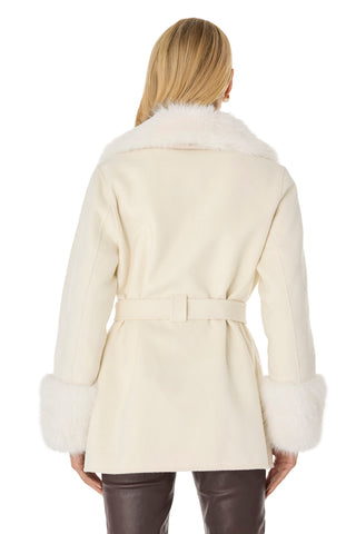 A person with long blonde hair, seen from behind, is wearing the Isabel Fur Trim Coat - White—a belted cream coat with fluffy white fur trim collar and cuffs—paired with dark pants.
