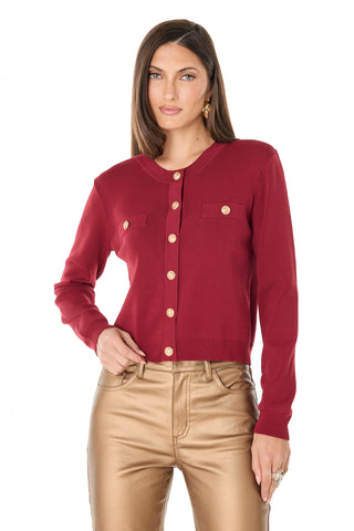 A woman wears the Loretta Knit Cardigan Top in merlot, featuring long sleeves, padded shoulders, two chest pockets, and gold dome buttons, paired with high-waisted metallic gold pants against a plain white background.