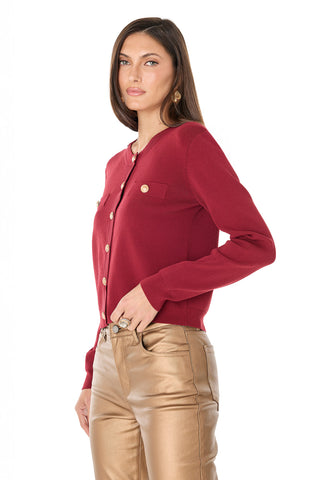 A woman with long brown hair wears the Loretta Knit Cardigan Top in merlot, featuring gold dome buttons, paired with tan leather pants against a white background.