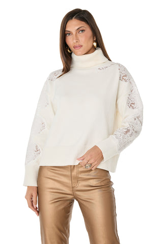 A woman models the Camille Lace Turtleneck Sweater - Pearl, featuring ultra-soft knit lace and delicate sleeve accents, paired with gold pants. She accessorizes with gold earrings and a statement ring against a white backdrop.