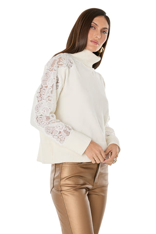 A woman wears the Camille Lace Turtleneck Sweater in Pearl with shiny bronze pants, standing with her hands together and gazing to the side against a white background.