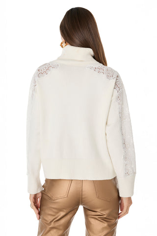 A woman with long brown hair models the Camille Lace Turtleneck Sweater in Pearl, showcasing intricate lace on the shoulders and sleeves. She pairs it with shiny bronze pants and faces away from the camera against a white background.