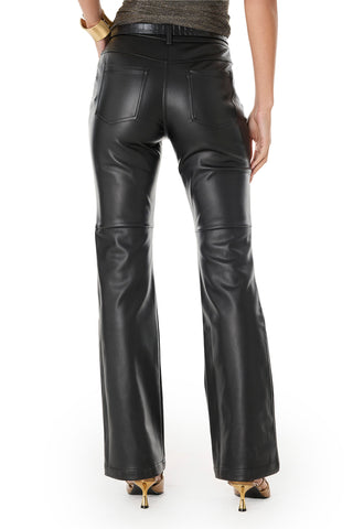 A person faces away wearing the Jackie Leather Pant - Black, a high-rise faux leather style with back pockets, a slight flare at the bottom, and paired with heeled shoes. Their left hand rests on their hip.