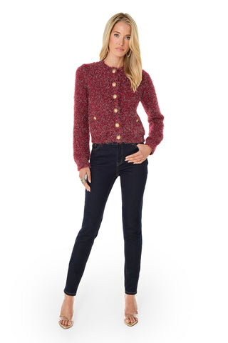 A woman faces forward against a plain white background, wearing the Joana Knit Cardigan - Merlot with gold dome buttons, paired with dark skinny jeans and beige heeled sandals.
