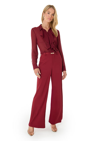 A woman poses confidently in the Georgia Semi-Sheer Blouse - Burgundy, a ruffled statement piece, styled with matching wide-leg trousers, gold earrings, rings, and open-toe heels against a plain white background.