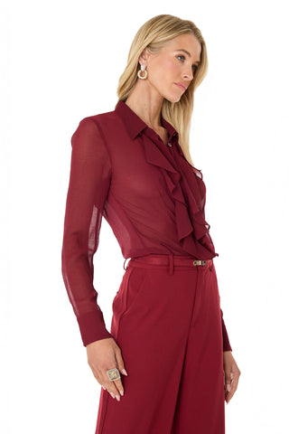 A woman with long blonde hair wears the Georgia Semi-Sheer Blouse in burgundy with front ruffles and matching high-waisted trousers, accessorized with gold hoop earrings and a large ring, posing against a white background.