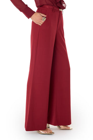 A person is shown from the waist down wearing the Martina Belted Trouser in Merlot—a high-waisted, wide-leg pant with a built-in belt—paired with gold pointed-toe heels, set against a white background.