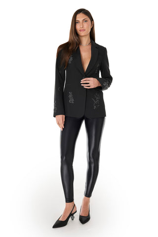 A woman with long hair stands against a white background wearing the London Floral Sequin Blazer in black, paired with shiny black leggings, pointed black heels, and a silver necklace. Her expression is neutral.