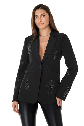 A woman with straight brown hair wears the London Floral Sequin Blazer in black, buttoned with no shirt underneath and paired with shiny black pants. She stands against a white background, looking confidently at the camera.