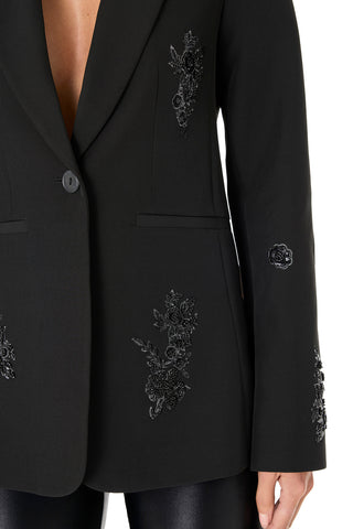 A person wears the London Floral Sequin Blazer - Black, a slim-fit piece with a deep neckline and black floral embroidery on the chest, sleeves, and front, paired with shiny black pants for a strikingly elegant look.