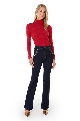 A woman faces forward in the slim-fitting Alina Rose Turtleneck Top - Red and dark high-waisted flare jeans with gold buttons, styled with blonde hair, gold earrings, and pointed-toe gold heels.