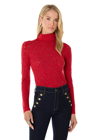 A woman with long blonde hair wears the Alina Rose Turtleneck Top - Red and dark blue high-waisted jeans with gold buttons. She faces forward, one hand resting at her side.