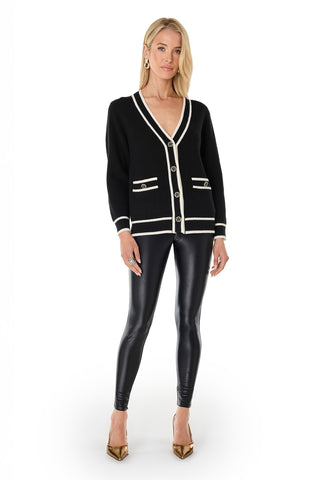 A woman with straight blonde hair and a neutral expression stands against a white background wearing the Devin Rose Contrast Cardigan - Black, shiny black leggings, gold pointed heels, and statement earrings.