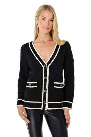 A woman with long blonde hair wears the Devin Rose Contrast Cardigan - Black, a v-neck style with white trim, metallic buttons, and two front pockets, paired with black leather pants. She accessorizes with hoop earrings and a ring against a white background.