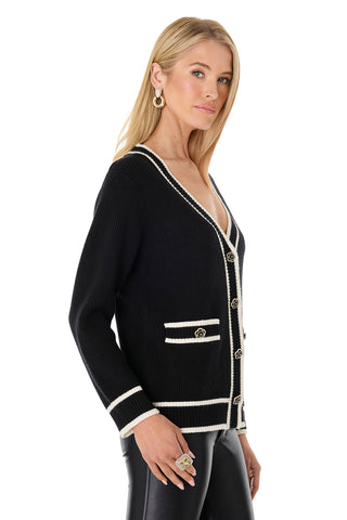 A woman with long blonde hair models the Devin Rose Contrast Cardigan in black, featuring white trim, gold buttons, and decorative pockets. She pairs it with black pants and poses sideways, looking at the camera against a white background.