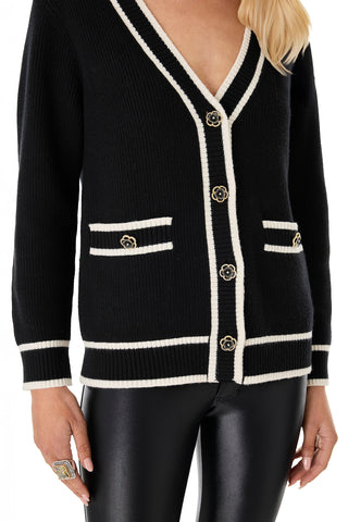 A woman wears the Devin Rose Contrast Cardigan - Black, featuring a v-neck, white trim, and rose buttons, paired with black leather pants. She has long blonde hair and accessorizes with a large statement ring.