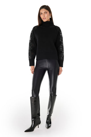 A woman with straight hair and a neutral expression stands against a white background, wearing the Camille Lace Turtleneck Sweater in black, paired with shiny black leather pants and knee-high heeled boots.