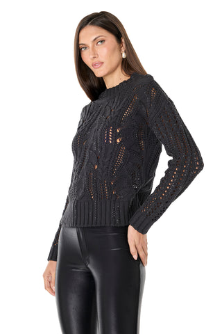 A woman wears the Elea Crystal Knit Sweater in black—a long-sleeve, open knit top with a textured pattern—paired with high-waisted shiny black pants, standing against a white background.