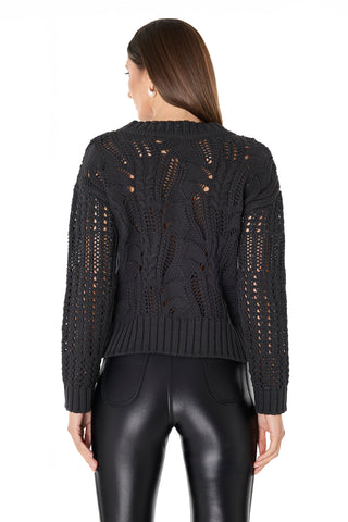 A woman with long brown hair, seen from behind, wears the Elea Crystal Knit Sweater in black with a leaf-like open knit pattern and shiny black leather-look pants against a white background.