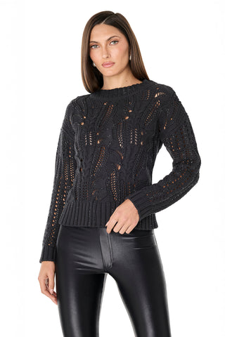 A woman with straight brown hair wears the Elea Crystal Knit Sweater in black, featuring an openwork texture, paired with shiny black leggings, standing against a plain white background.
