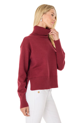 A woman with long blonde hair wears the Gia Ultra Soft Sweater in Merlot and white pants, standing against a white background, looking to the side with one hand touching her chin.