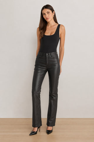 A woman wearing the Loren Bodysuit - Black, paired with black leather pants and black heels, stands against a plain light-colored wall. She looks at the camera with one hand in her pocket.