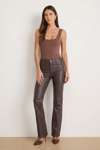 A woman stands against a plain wall wearing the Loren Bodysuit - Espresso, crafted from stretch fabric for a curve-hugging fit. She pairs it with dark brown leather pants and brown heels. Her long hair is styled simply, she wears minimal jewelry, and her expression is neutral.
